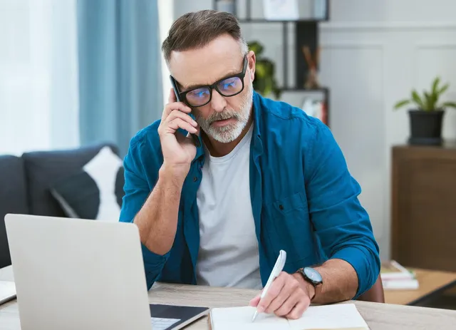 Mężczyzna w niebieskiej koszuli rozmawia przez telefon i robi notatki